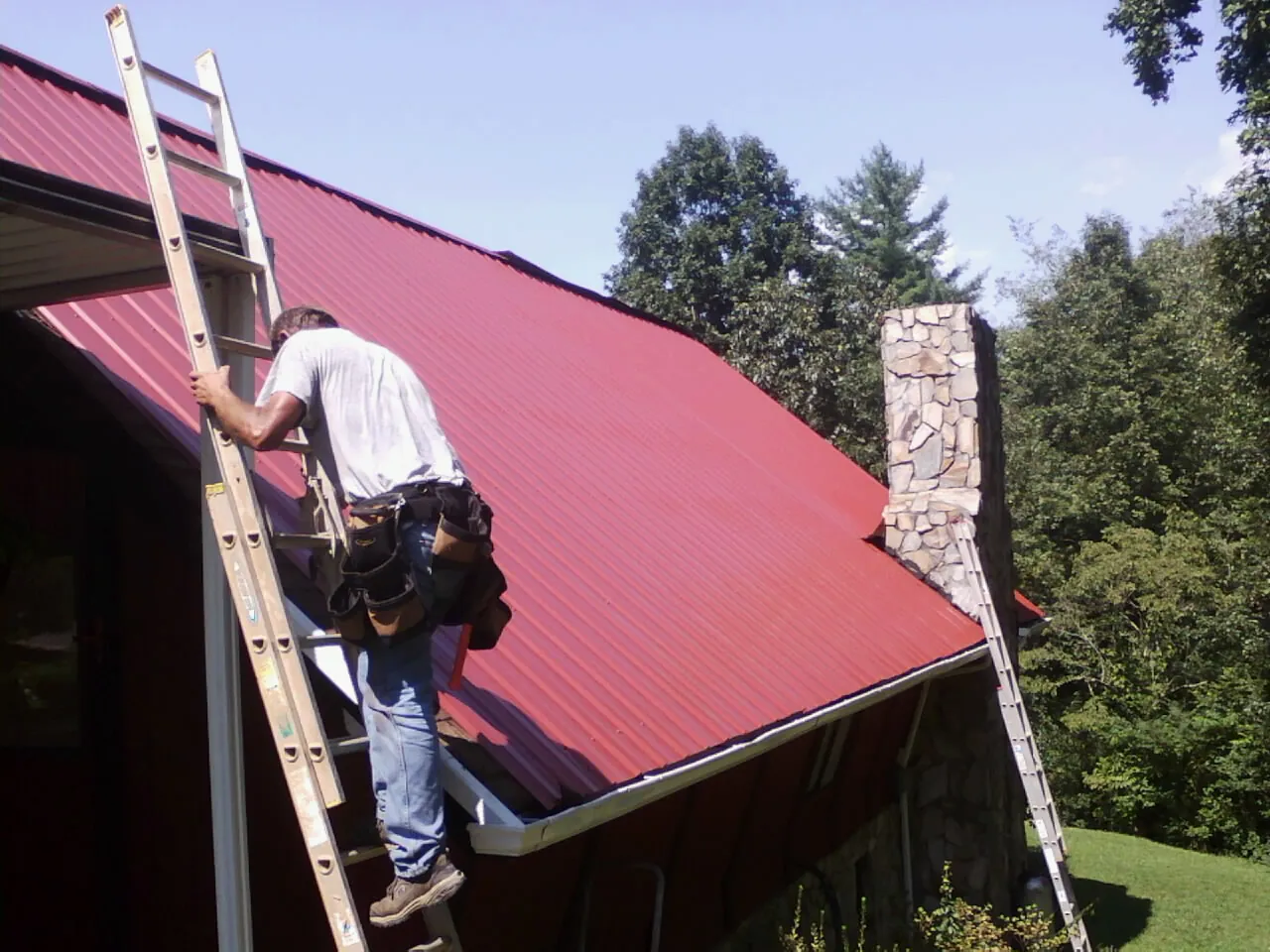 Roofer on a ladder beside a red metal roof and stone chimney — Soffit & Fascia Repair by Roofing Brothers in Valhalla